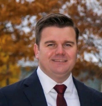 A professional headshot photo of Naim Jusic in a dark suit, white shirt and maroon tie. Fall foliage in the background.