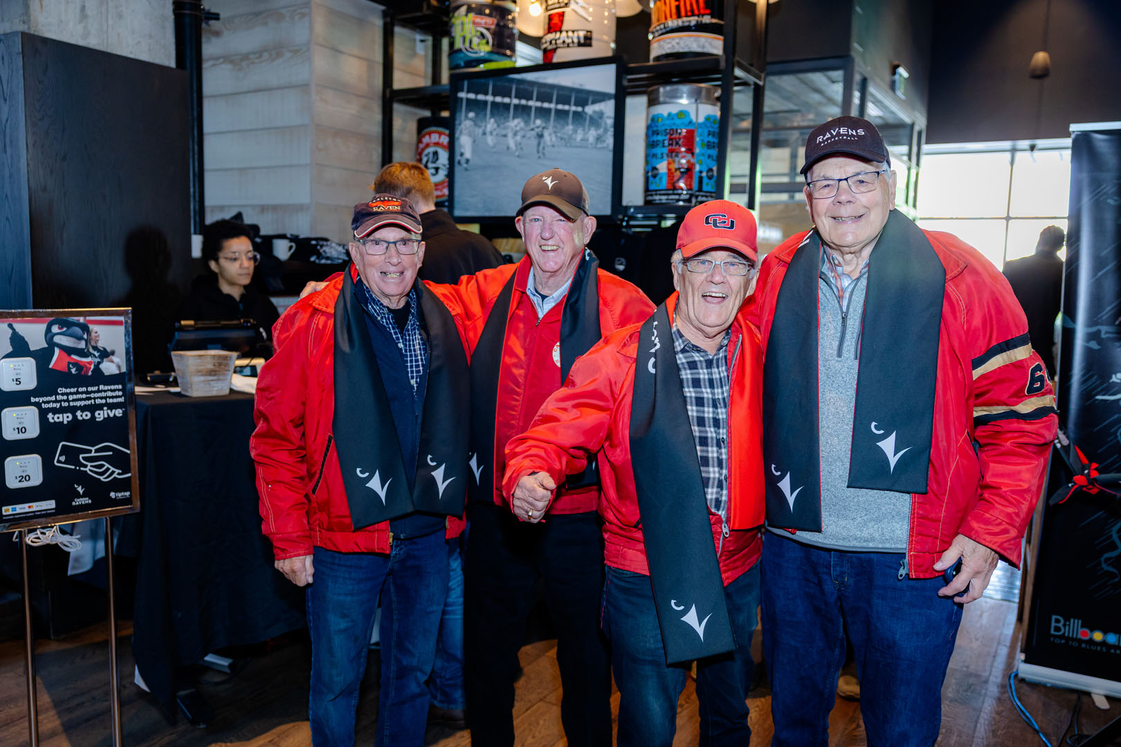 Four people smile wearing matching red jackets and Ravens scarves